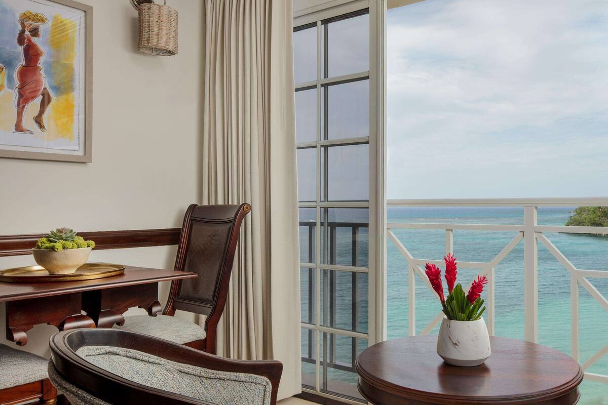 A room with a view of the ocean, a dining table with a bowl of fruit, a chair, and a vase with red flowers.