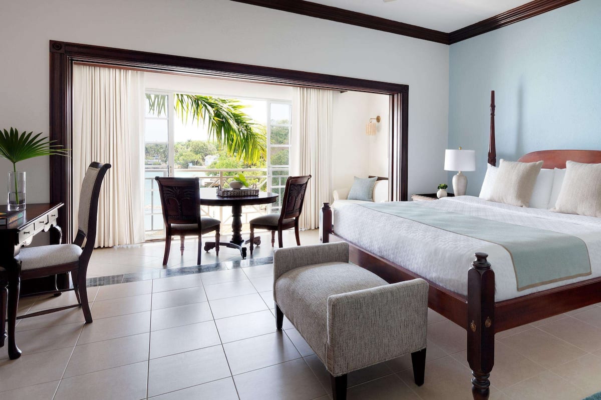 A hotel room with a large bed, a dining area with chairs, a small table with a plant, and a view of the outdoors through a sliding door.