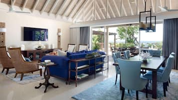 A spacious living area with a blue sofa, wicker chairs, a dining table, and a large TV.
