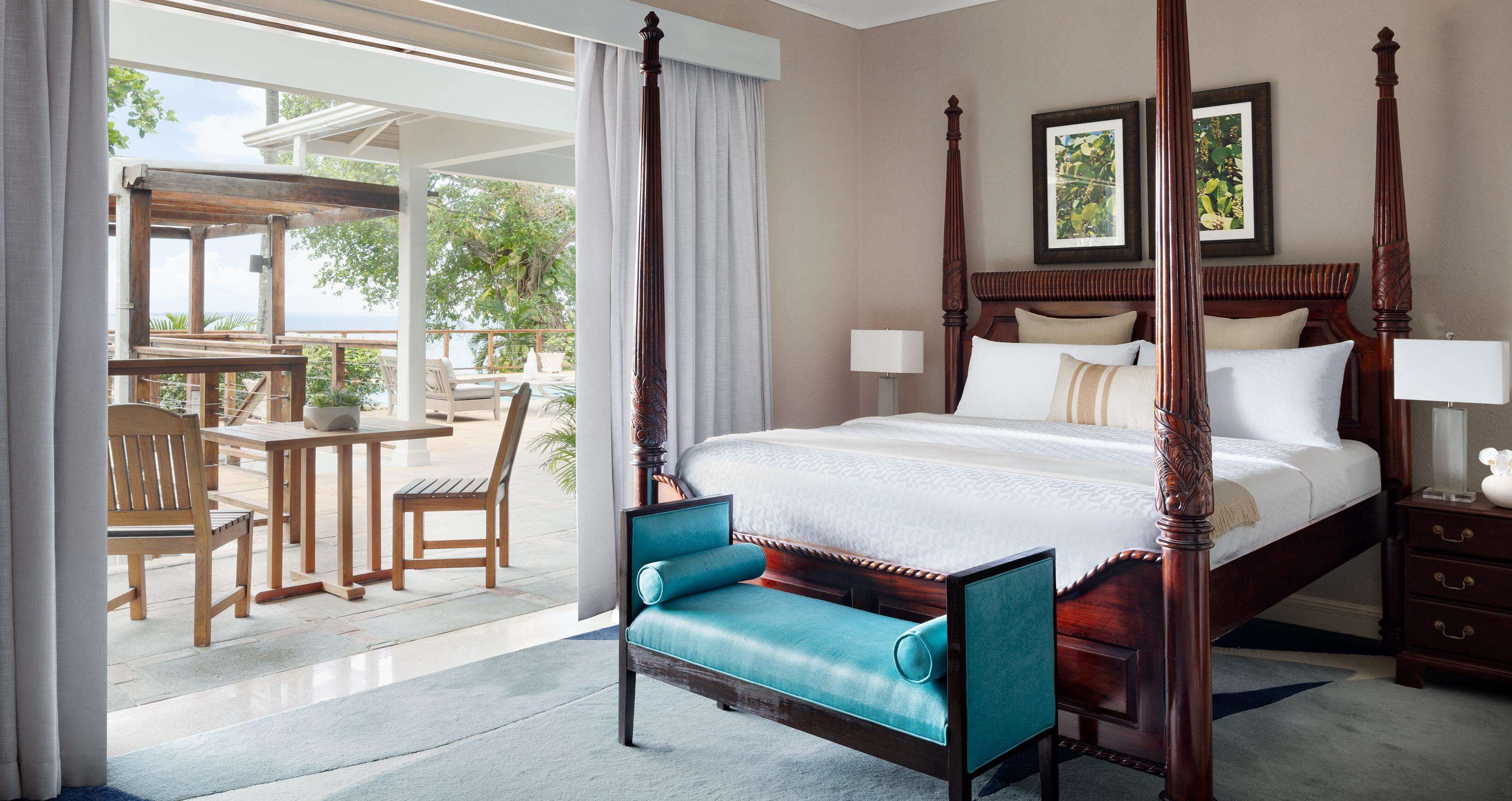 A four-poster bed with a wooden headboard, a teal upholstered chair, a wooden desk with a chair, and a view of the outdoors.