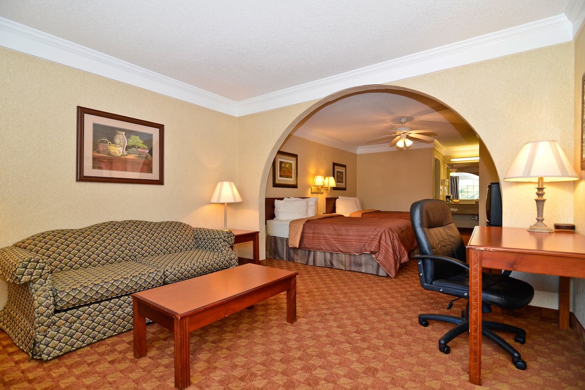 A hotel room with a sofa, a bed, a desk, and a chair. There is a ceiling fan, two wall-mounted lamps, and framed artwork on the wall.