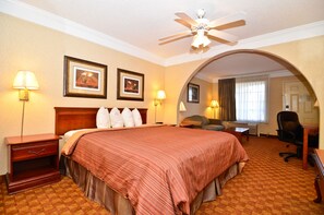 A hotel room with a large bed, two wall-mounted lamps, a ceiling fan, and an arched doorway leading to another room.