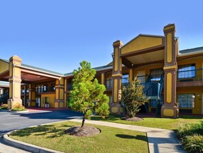 A multi-story hotel with a yellow facade, balconies, and a landscaped front yard with trees.