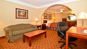 A hotel room with a sofa, a desk, a chair, a bed, and a bathroom visible through an archway. - Peach State Inn & Suites (Hawkinsville)