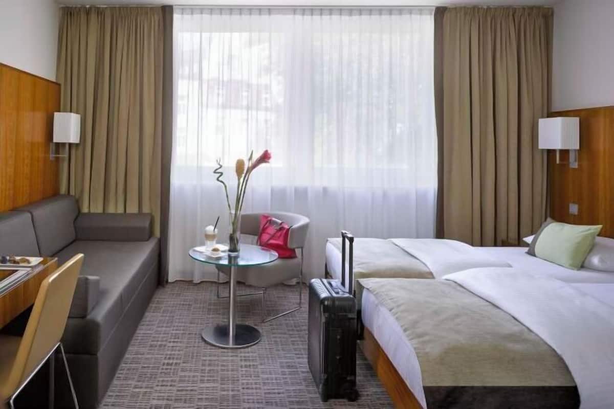 Habitación de hotel con cama, escritorio, silla, una pequeña mesa con un florero y una maleta.