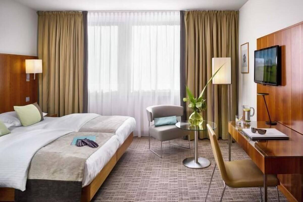 Una habitación de hotel con cama, escritorio, silla, televisor y ventana con cortinas.