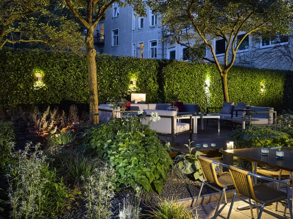 An outdoor patio area with seating and tables, surrounded by lush greenery and a hedge wall. - K+K Hotel am Harras (Munich)