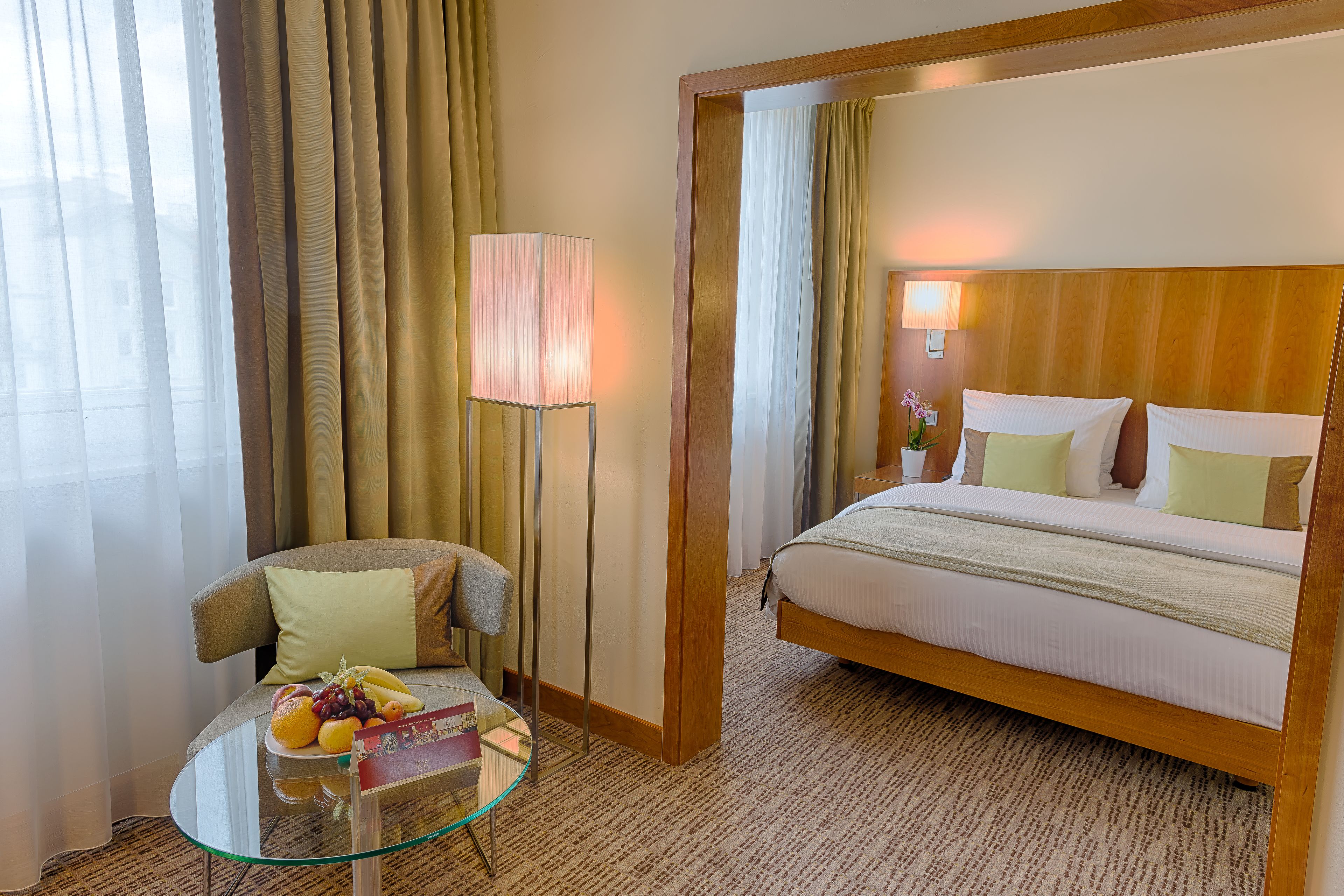 A hotel room with a bed, a chair, a glass table with fruits, and a lamp.