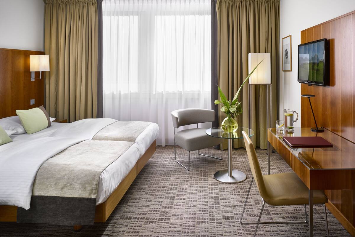 Habitación de hotel con cama, escritorio, silla, televisor y ventana con cortinas.