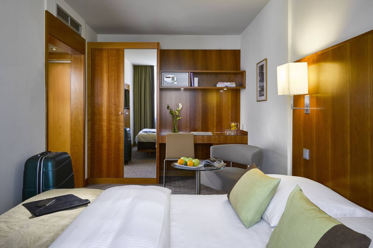 Habitación de hotel con cama, escritorio, silla, pequeña mesa con frutas y una maleta.