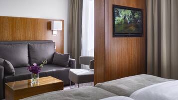 Ein Hotelzimmer mit grauem Sofa, einem Holztischchen mit Blumen in einer Vase, einem kleinen Sessel, einem an einer Holzwand montierten Fernseher und zwei Betten mit grauer Bettwäsche.