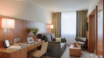 Habitación de hotel con escritorio de madera, computadora portátil, silla, sofá y una mesita con fruta y bebidas.