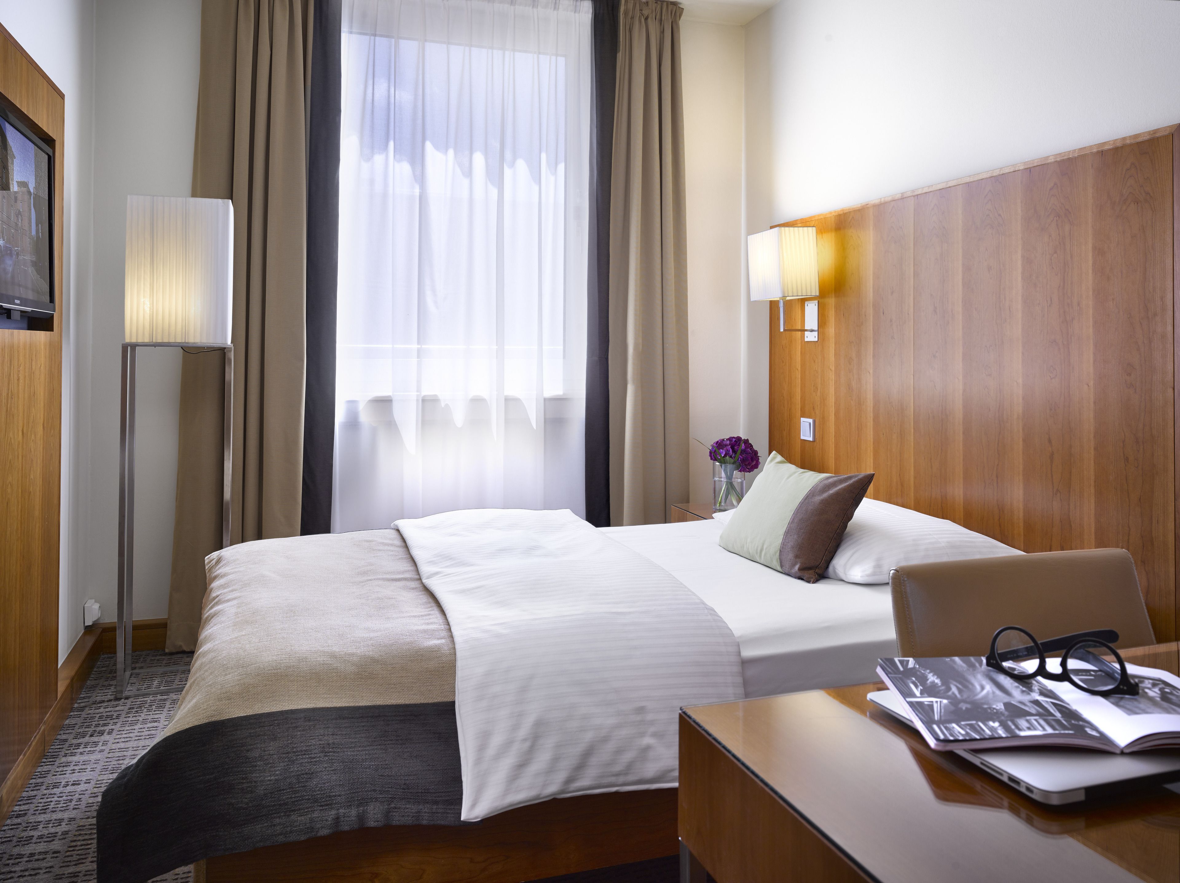A hotel room with a bed, bedside table, lamp, and a view of the outdoors through a window.