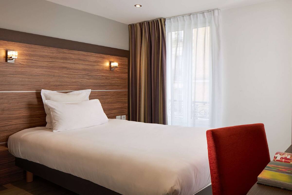 Une chambre d’hôtel avec une tête de lit en bois, un lit recouvert de draps blancs, une chaise rouge et une grande fenêtre avec des rideaux.