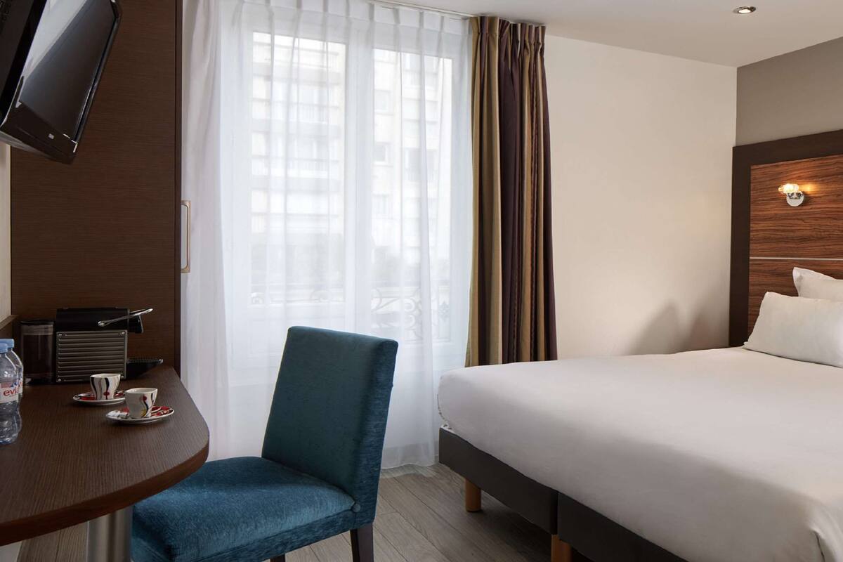 Une chambre d’hôtel comprenant un lit, un bureau avec une chaise, une télévision et une fenêtre avec des rideaux.