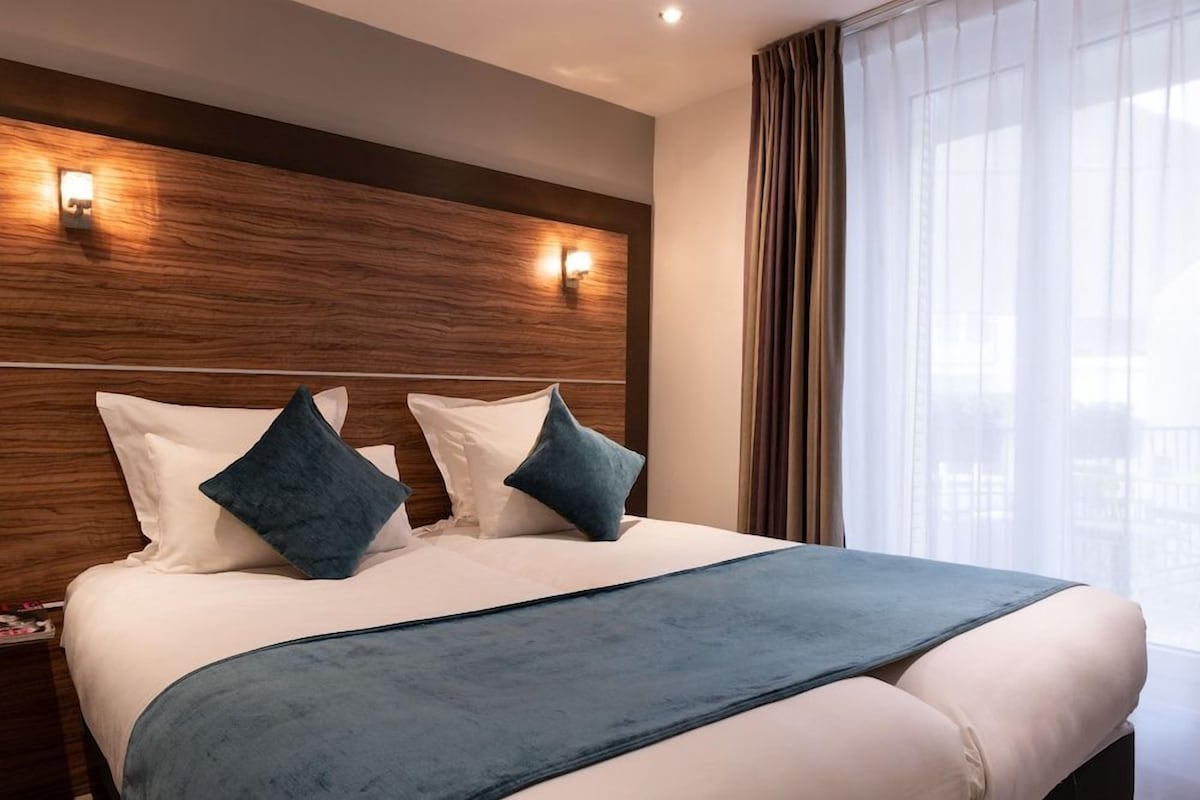 Een hotelkamer met een groot bed, twee wandlampen, een raam met gordijnen en een houten hoofdeinde.