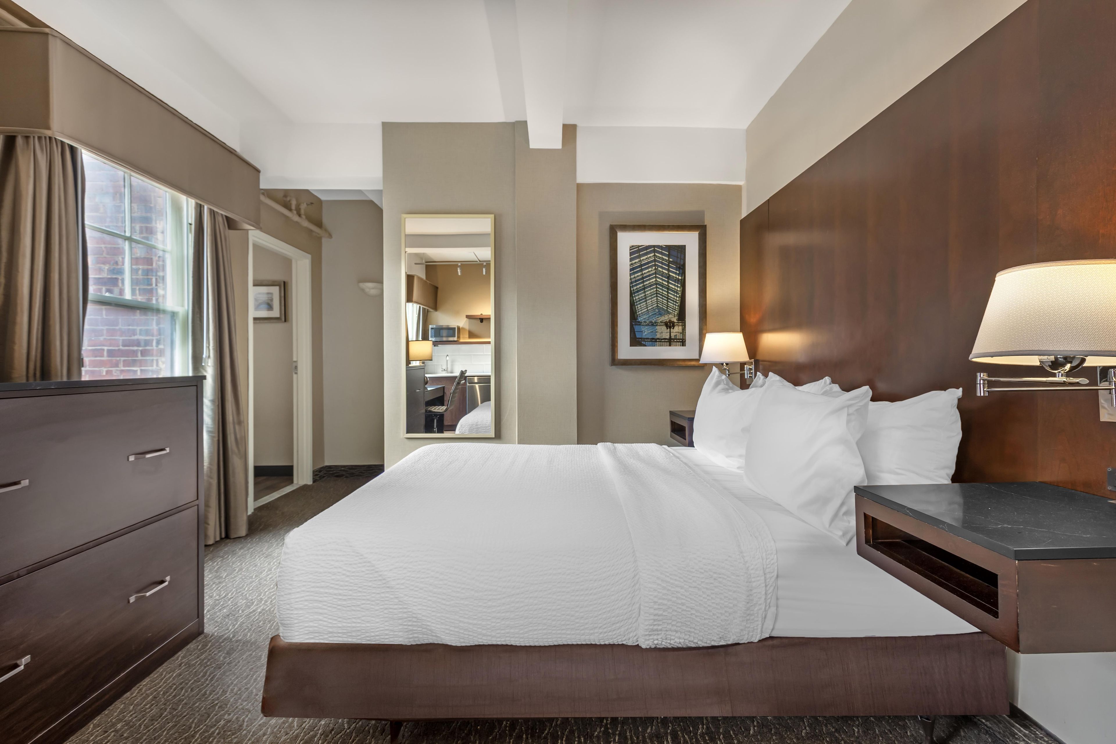 A hotel room with a bed, bedside table, lamp, and a view of the hotel's interior.