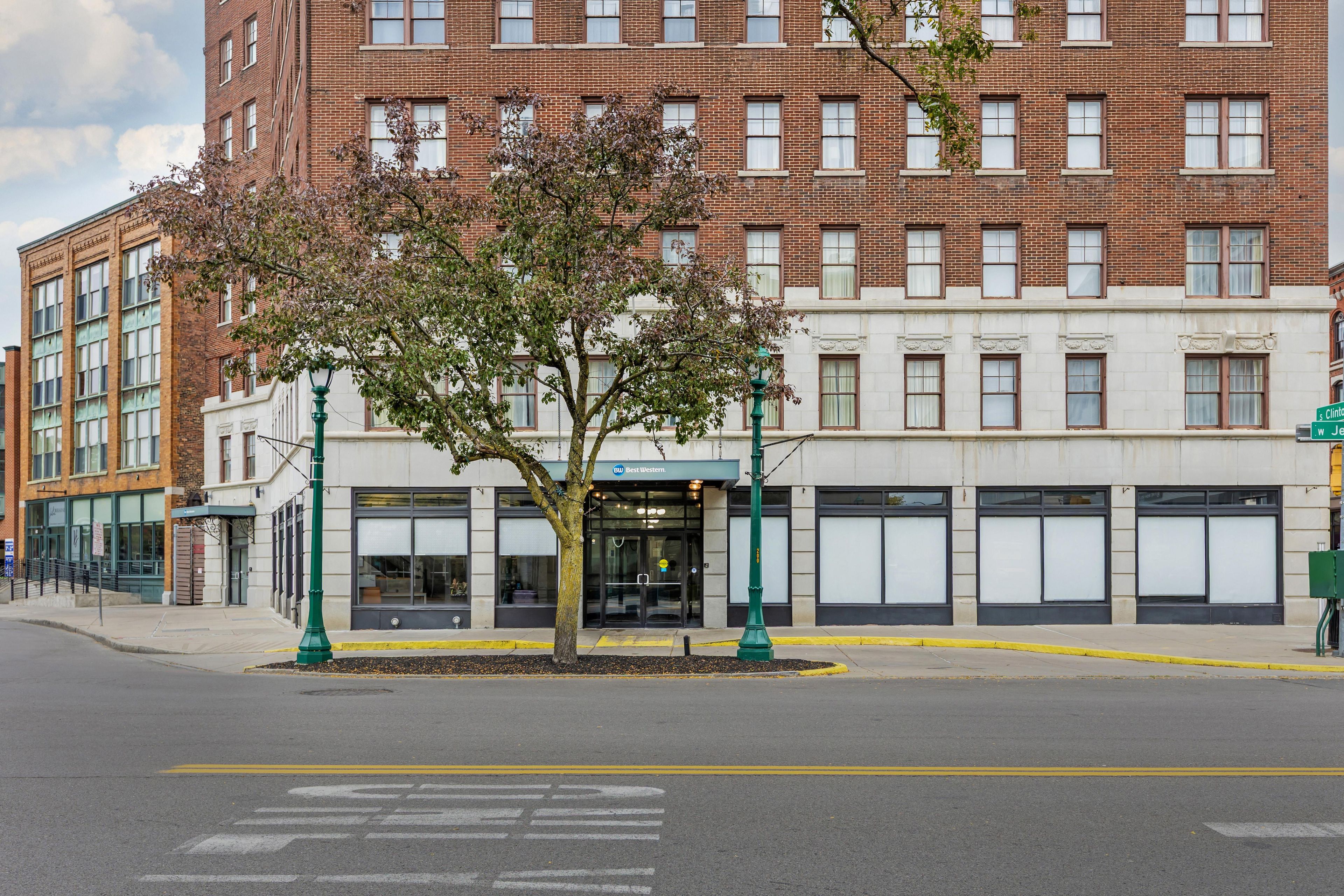 A street view with a building featuring a brick facade, a tree in the foreground, and storefronts with closed shutters.