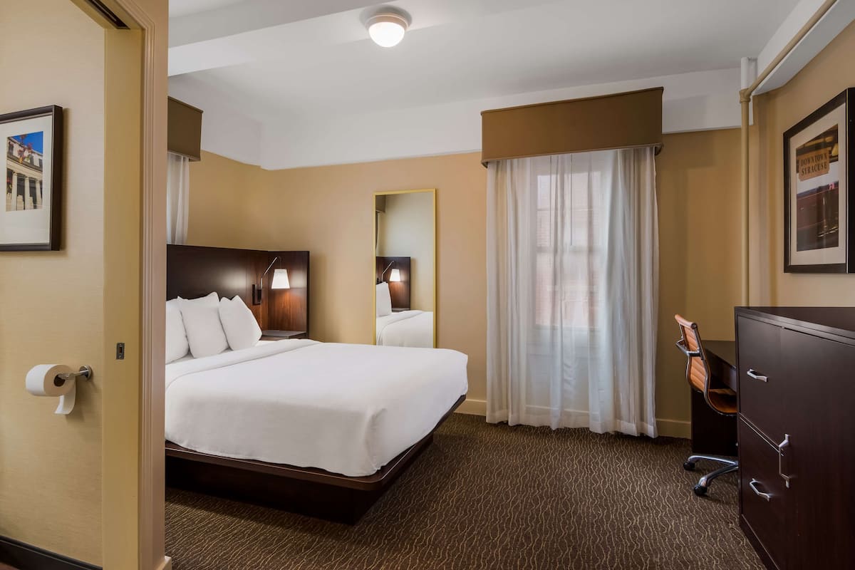 A hotel room with a large bed, a desk with a chair, a dresser, a mirror, and a framed picture on the wall.