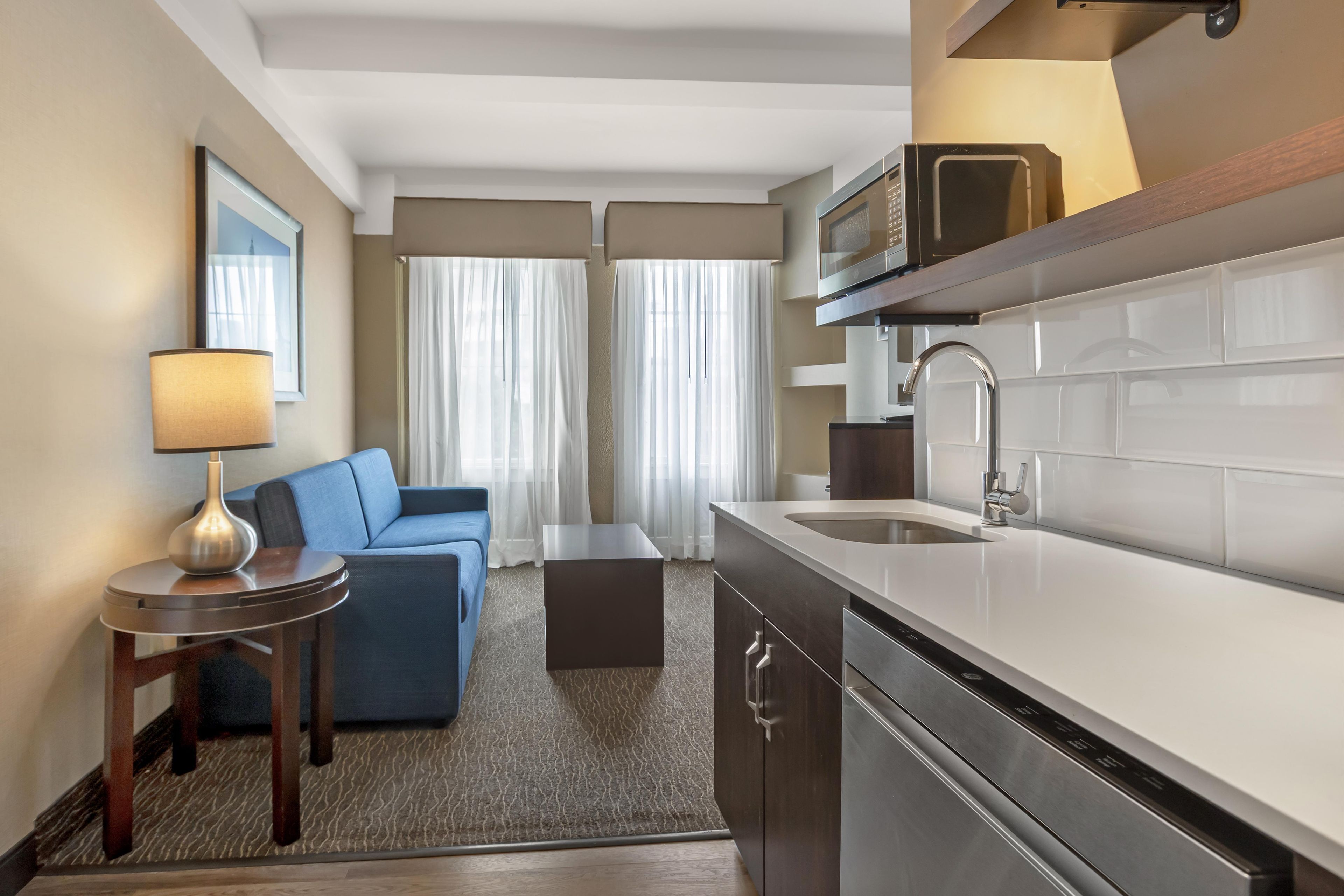 A compact hotel room with a kitchenette, a sofa, a small table, and a lamp.