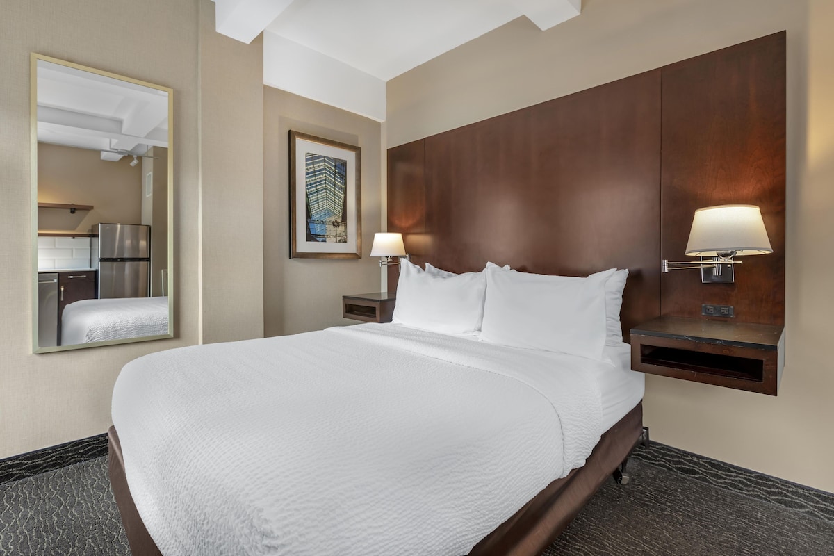 Habitación de hotel con una cama grande, una mesita de noche con lámpara, un espejo y un cuadro en la pared.