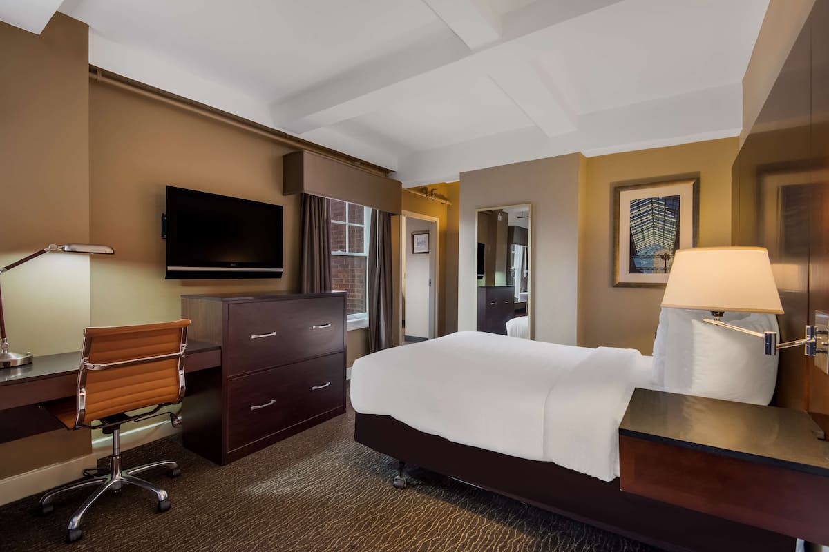 A hotel room with a bed, desk, chair, TV, wardrobe, and a painting on the wall.