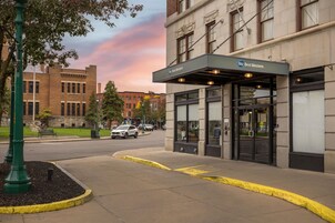 Entrada de un hotel con un área exterior cubierta, vista de la calle con coches estacionados y un edificio con fachada de ladrillo al fondo.