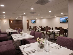 Un comedor de hotel con varias mesas, sillas y una pantalla de televisión que muestra una vista panorámica.
