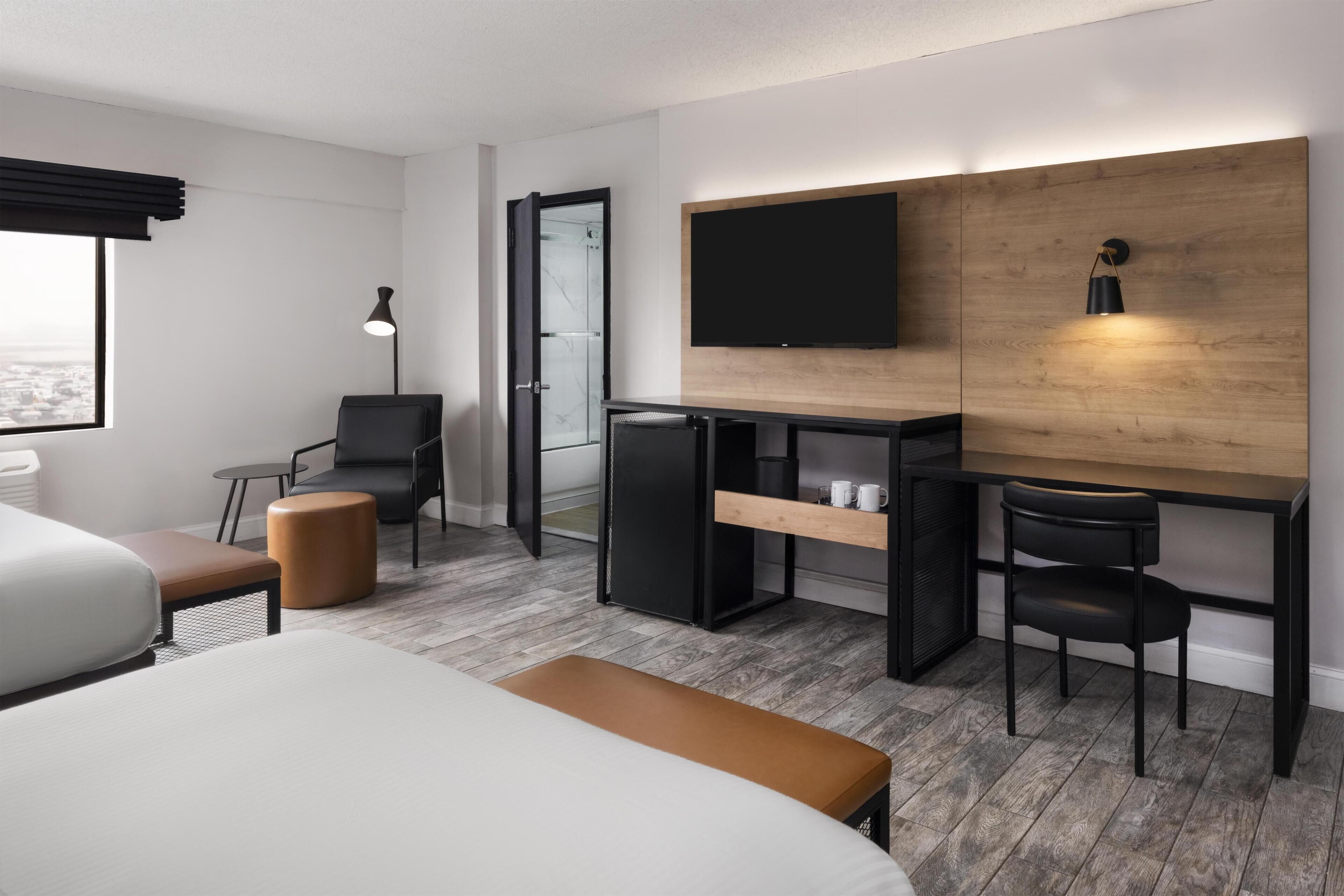 A modern hotel room with a bed, desk, chairs, a TV, and a view of the city.