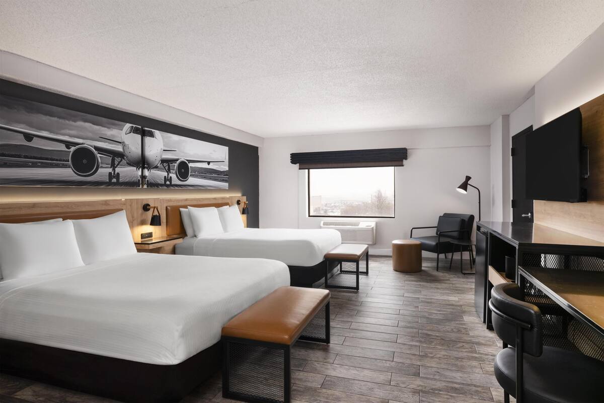 A hotel room with two beds, a desk, a chair, a TV, and a large window.
