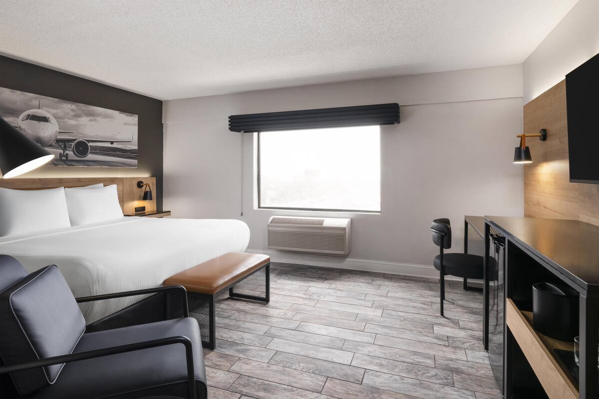 A hotel room with a bed, a desk, a chair, a bench, a window with blinds, an air conditioning unit, and a wall-mounted TV.