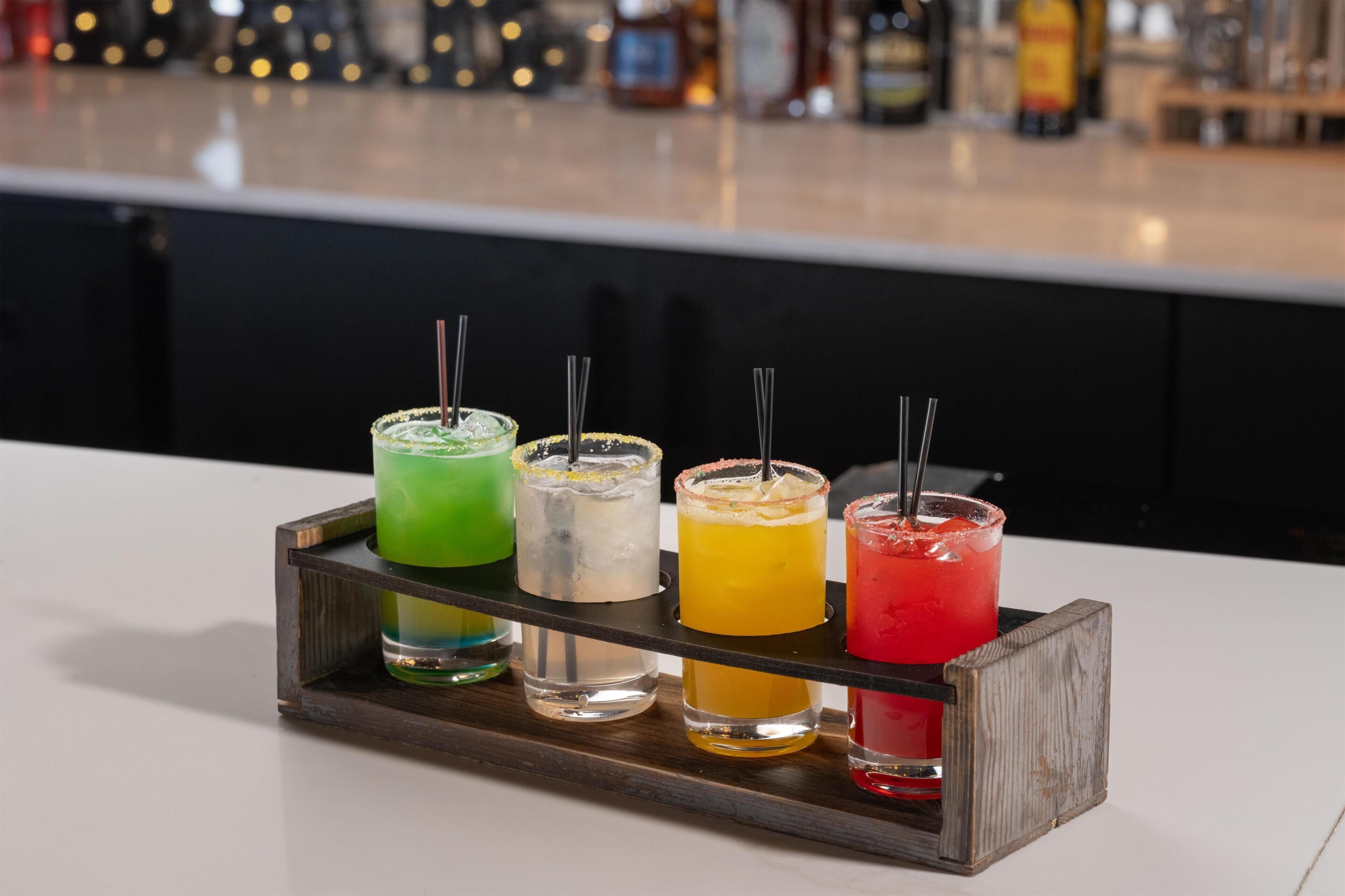Un área de bar con una bandeja de madera que sostiene cuatro vasos de bebidas de diferentes colores.