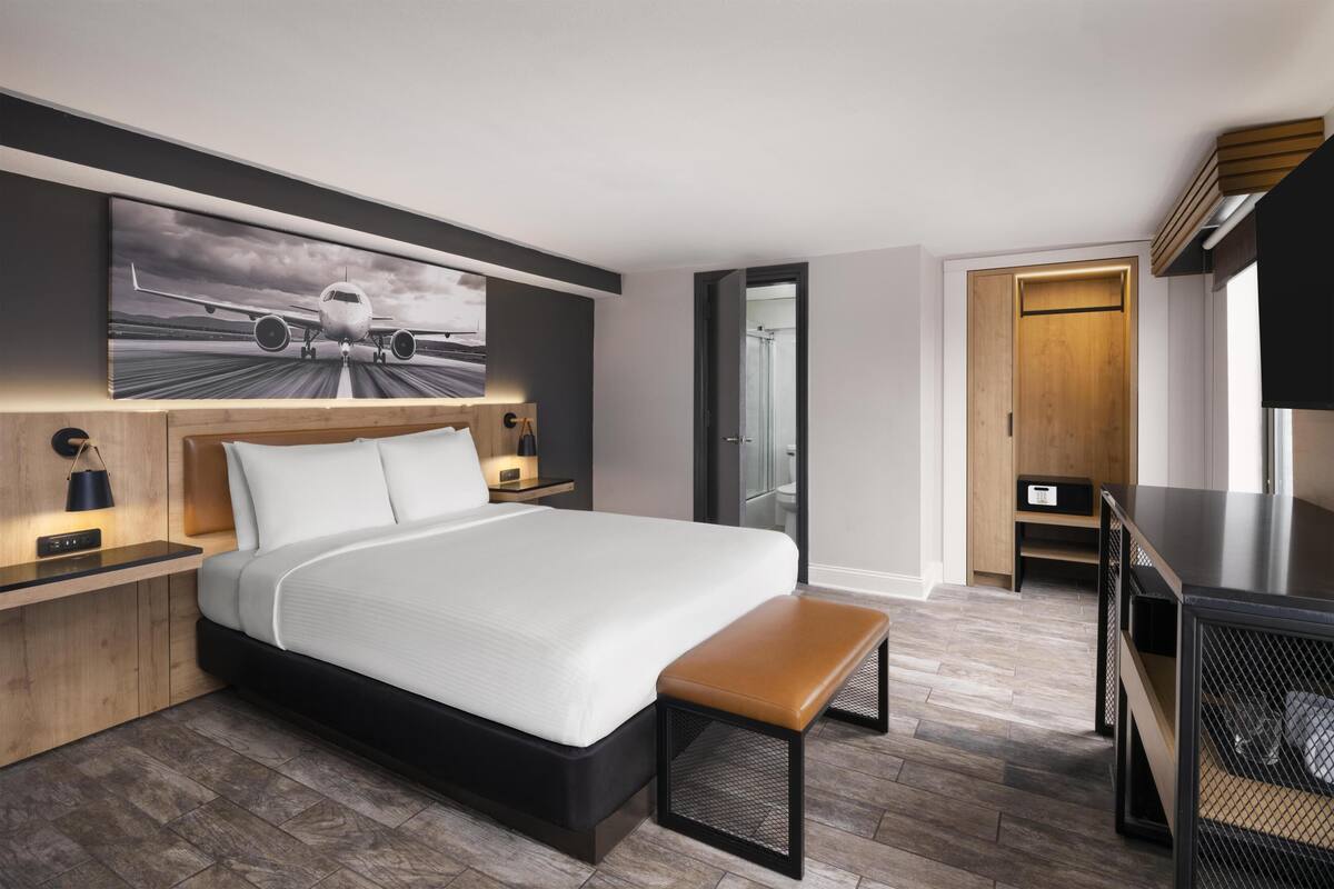 A modern hotel room with a large bed, a bench, a desk, and a large window.