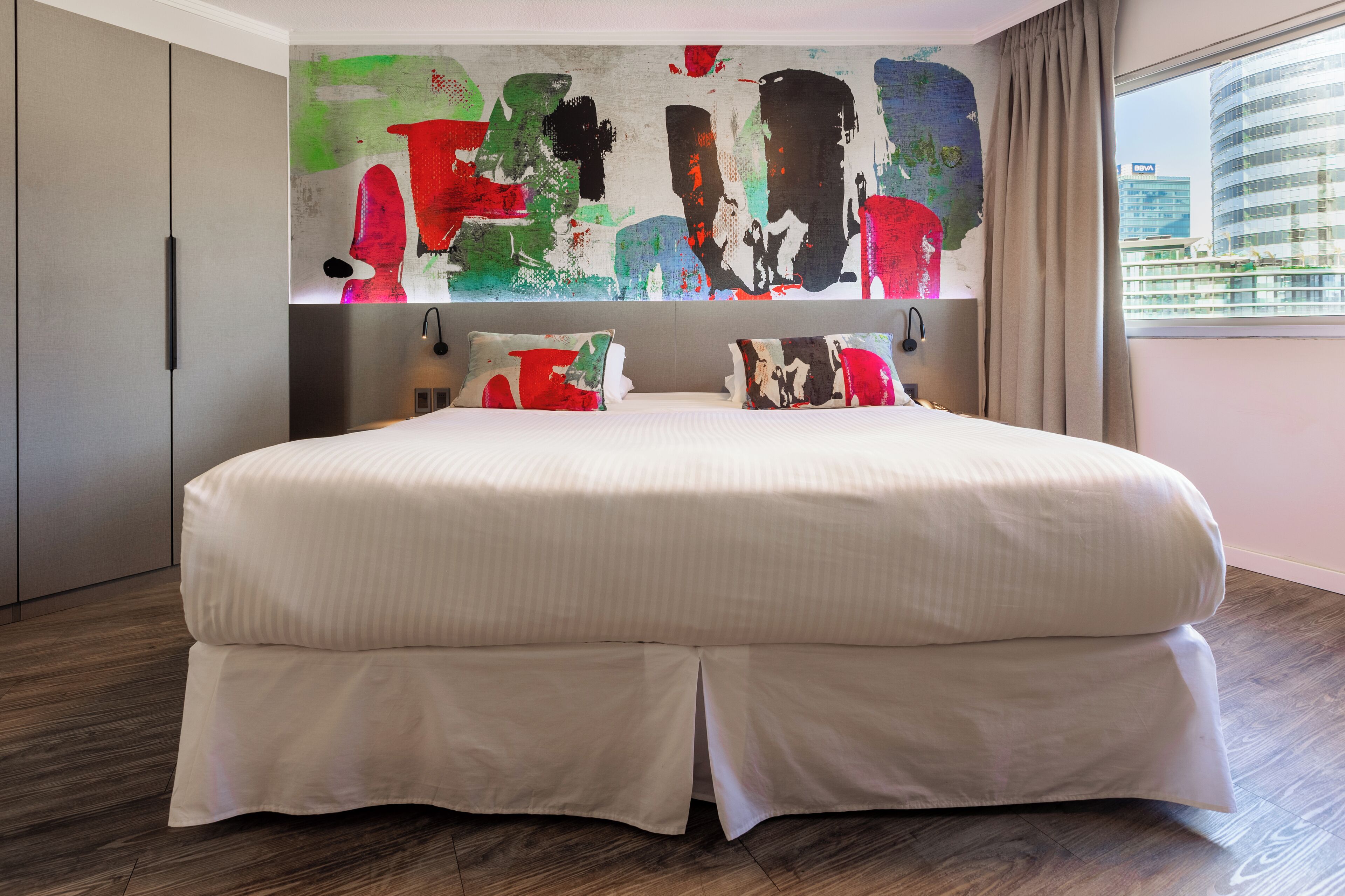 Ein großes Bett mit weißer Bettdecke und zwei Kissen, ein modernes abstraktes Bild an der Wand und ein Blick auf eine Stadtansicht durch ein Fenster.