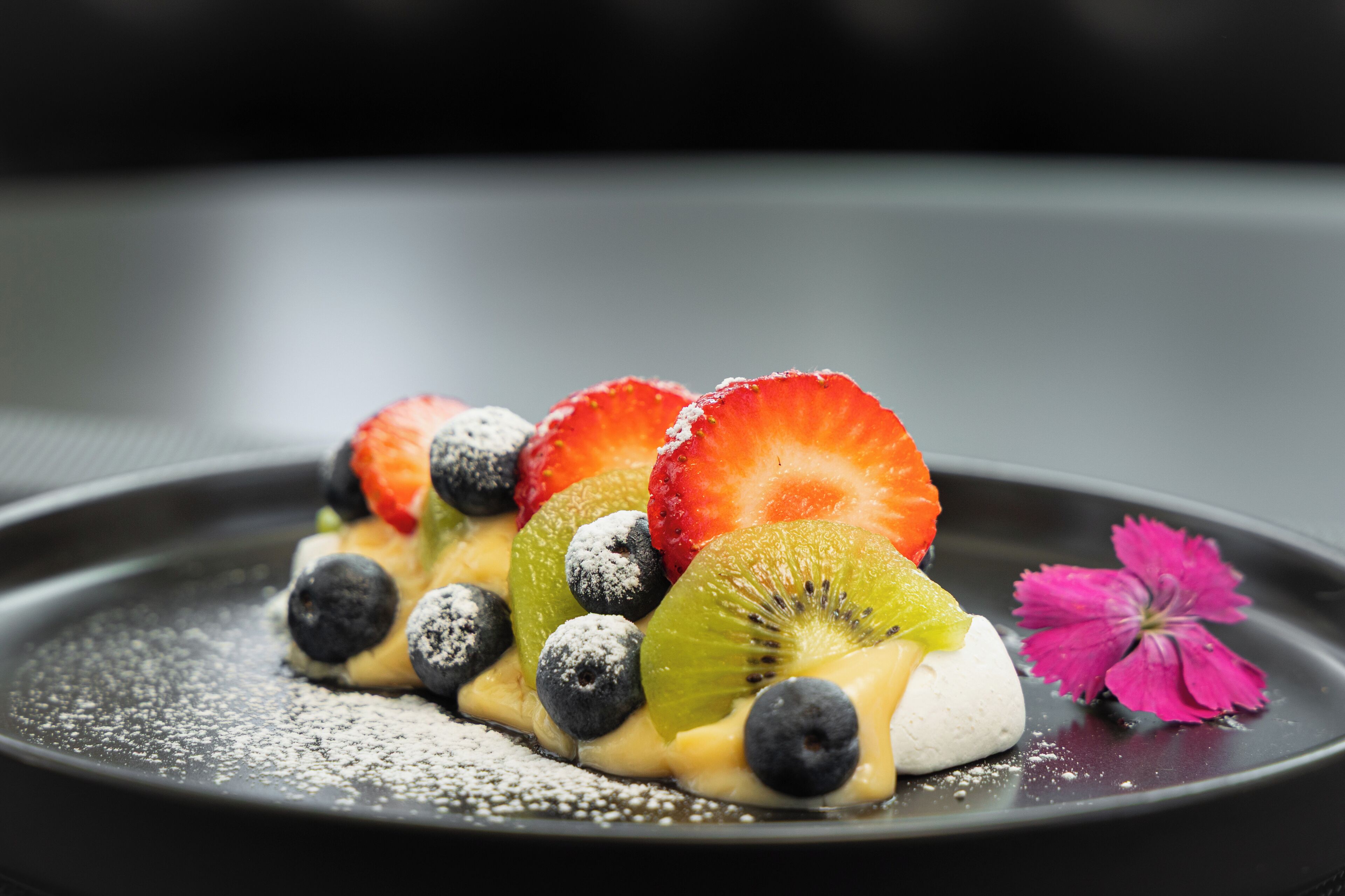 Postre con fresas, arándanos, kiwi y una flor rosa sobre un plato negro.