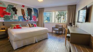 Hotellihuone, jossa on suuri sänky, työpöytä, tuoli, televisio ja ikkuna, josta avautuu näkymä palmuille ja rakennuksille.