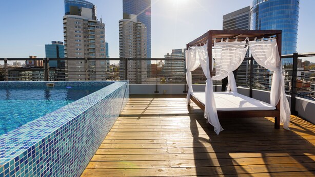 A rooftop pool area with a wooden deck, a bed with white curtains, and a city skyline in the background. - Palladium Business Hotel (Montevideo)