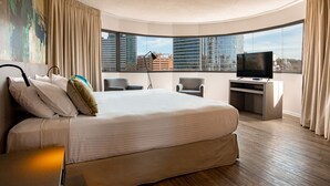 A modern hotel room with a large bed, a desk with a TV, and a view of the cityscape through large windows. - Palladium Business Hotel (Montevideo)
