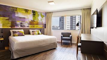 Una habitación de hotel moderna con una cama grande, un escritorio, una silla, un amplio ventanal con vistas a la ciudad y un cuadro abstracto de colores en la pared.