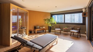 A modern room with a wooden floor, a massage table, a large window with a city view, a potted plant, and a small table with a chair. - Palladium Business Hotel (Montevideo)