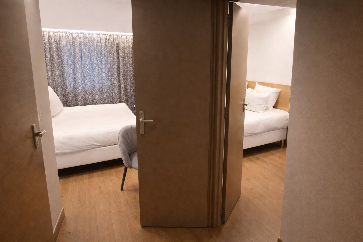A hotel room with a wooden floor, a bed with white bedding, a chair, and a door.