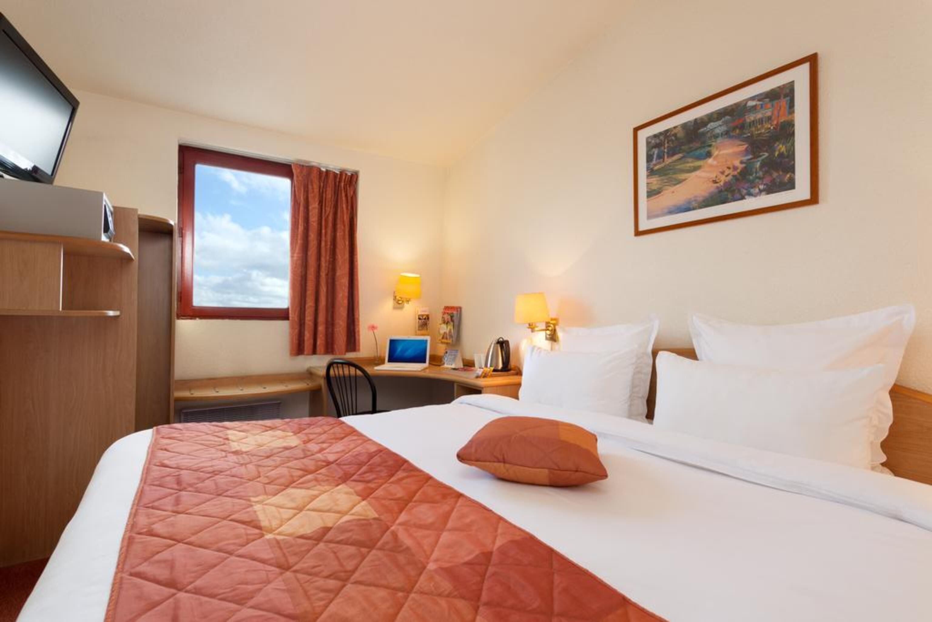 A hotel room with a bed, a desk, a chair, a TV, and a painting on the wall.