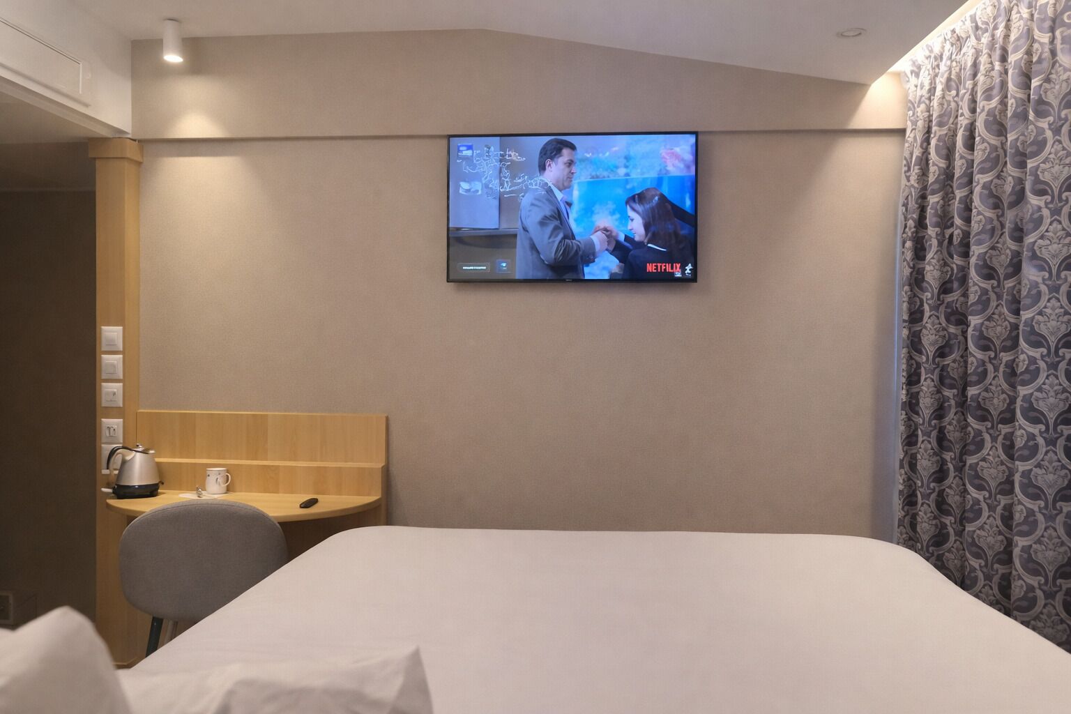 Habitación de hotel con cama, escritorio con hervidor y taza, televisión montada en la pared y cortina estampada.