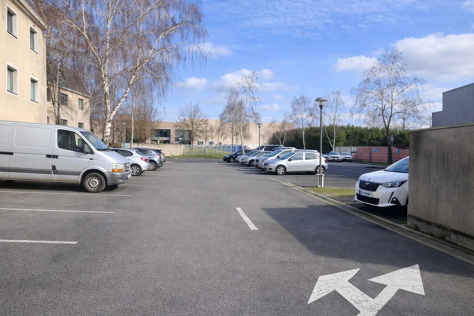 A parking lot with several cars and a van, lined with buildings and leafless trees.