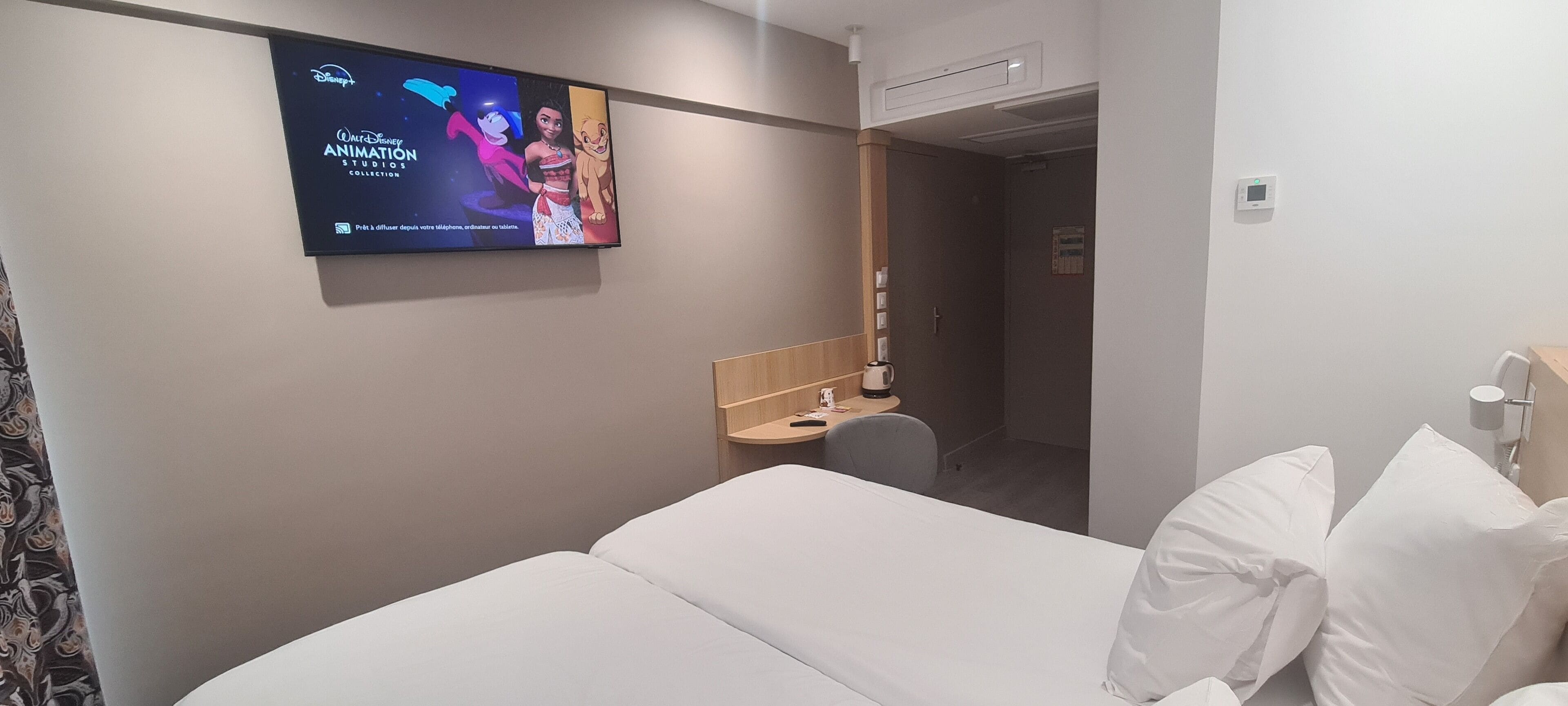 A hotel room with a bed, a desk, a TV displaying Disney's 'Walt Disney Animation Studio Collection', and a door leading to another room.