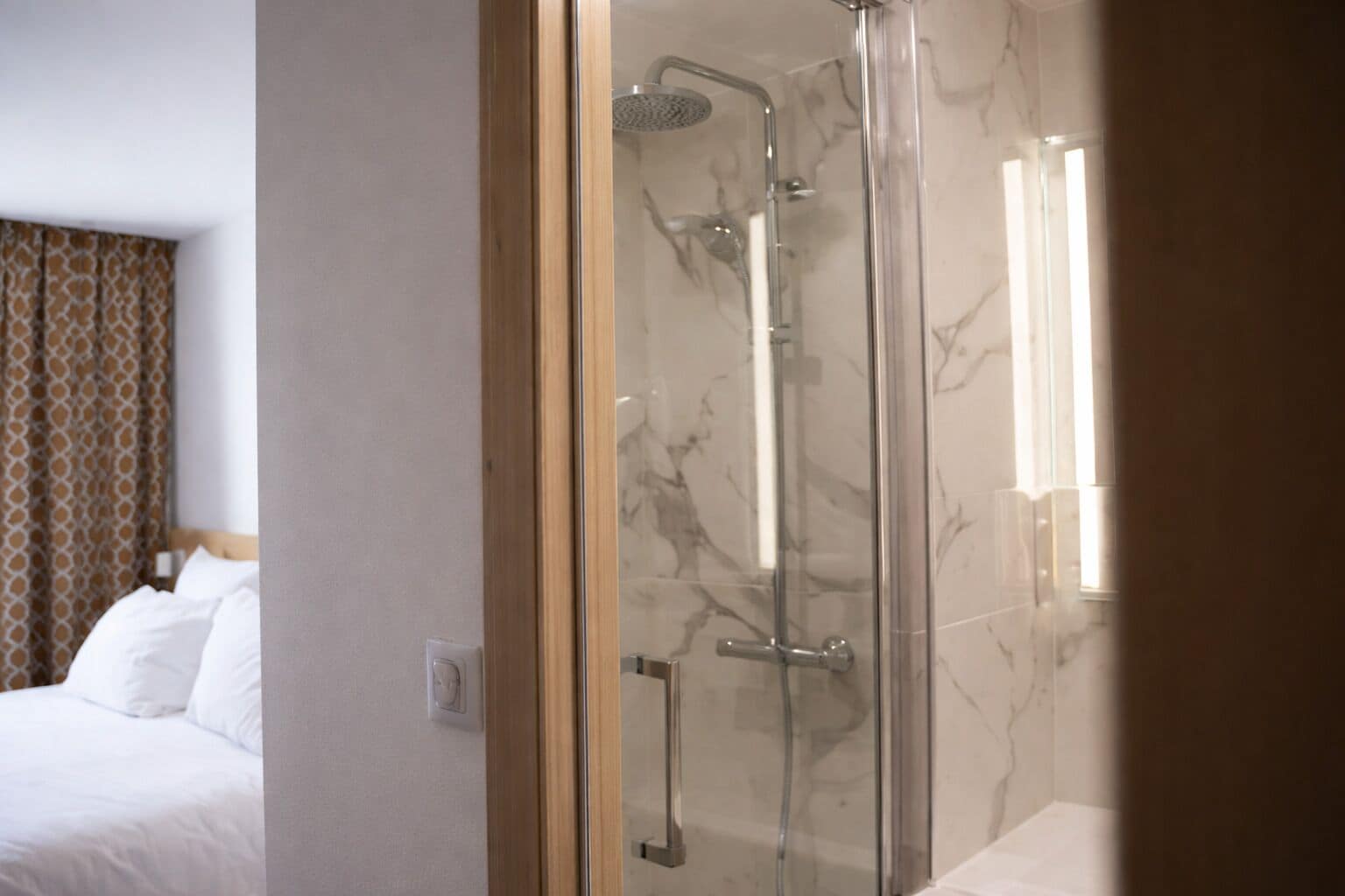 A hotel room with a glass-enclosed shower, a bed with white linens, and a patterned curtain.