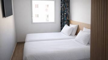 Quarto de hotel com duas camas, janela com cortinas e uma televisão de tela plana fixada na parede.