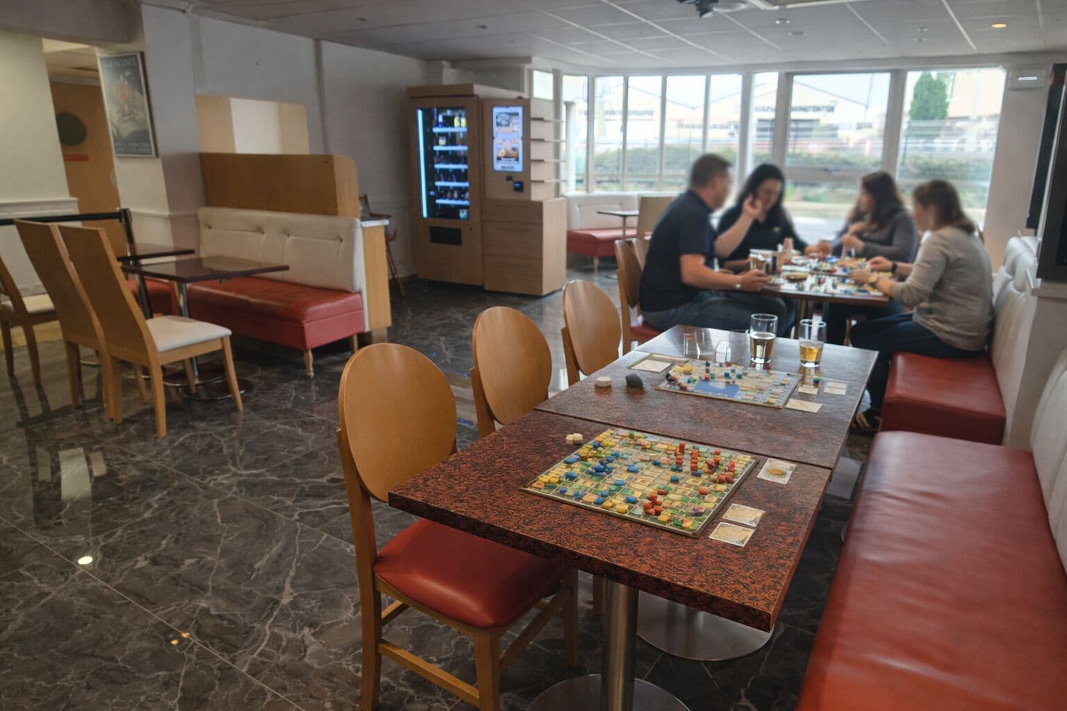A restaurant with marble flooring, wooden chairs, and tables. There are people dining and a vending machine in the background.