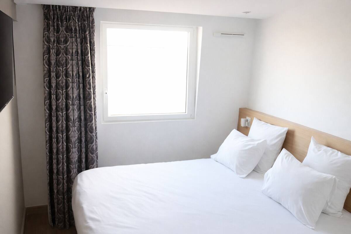 Ein Hotelzimmer mit Bett, weißen Vorhängen, einem Fenster und einer Wand-Klimaanlage.
