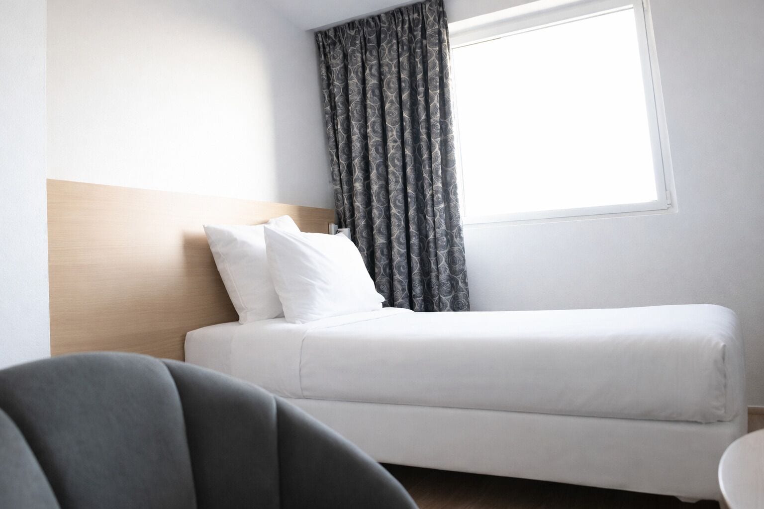 Une chambre d’hôtel avec un lit simple, une tête de lit, une fenêtre avec des rideaux et une vue partielle sur une chaise.